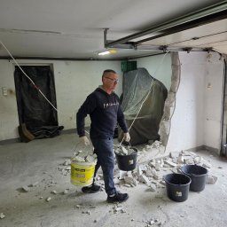 3Miasto-Hol Piotr Szpreniec - Mężczyzna w okularach usuwa gruz po wyburzeniu ściany w garażu. Widoczne wiadra z resztkami, fragment ściany i zabezpieczone folią elementy. Praca w toku.