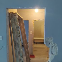 3Miasto-Hol Piotr Szpreniec - Wyburzona ściana z widoczną futryną i fragmentem łazienki w tle. Ślady po kuciu, surowy stan po demontażu. Czerwona zmiotka oparta o ścianę.