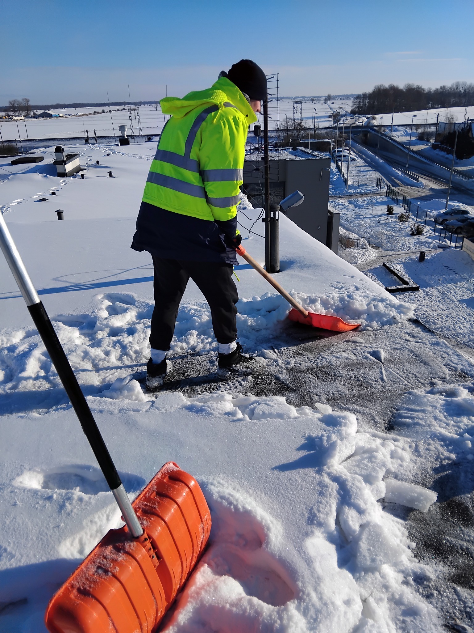 Pracownik w odblaskowej kurtce odśnieża płaski dach budynku za pomocą pomarańczowej łopaty. W tle widoczny krajobraz miejski pod błękitnym niebem.