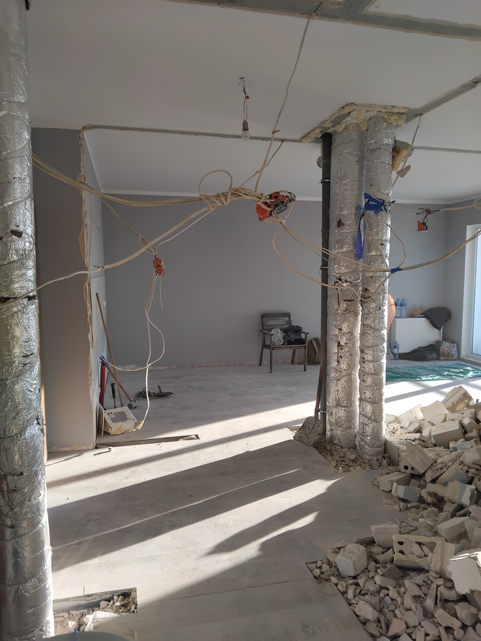 Wnętrze w trakcie demontażu: odsłonięte przewody, zabezpieczone rury, gruz. Widoczne narzędzia i fragmenty ścian. Jasne światło podkreśla surowość stanu obecnego.