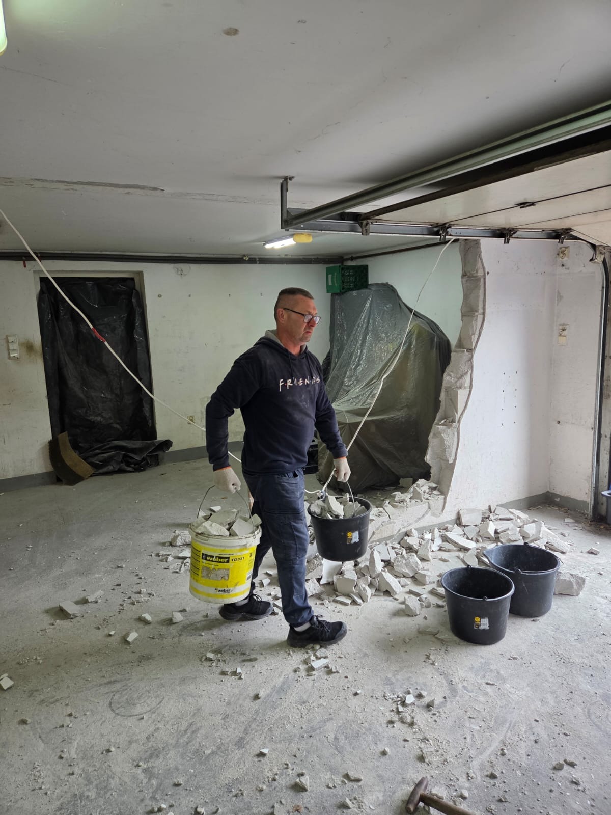 Mężczyzna w okularach usuwa gruz po wyburzeniu ściany w garażu. Widoczne wiadra z resztkami, fragment ściany i zabezpieczone folią elementy. Praca w toku.