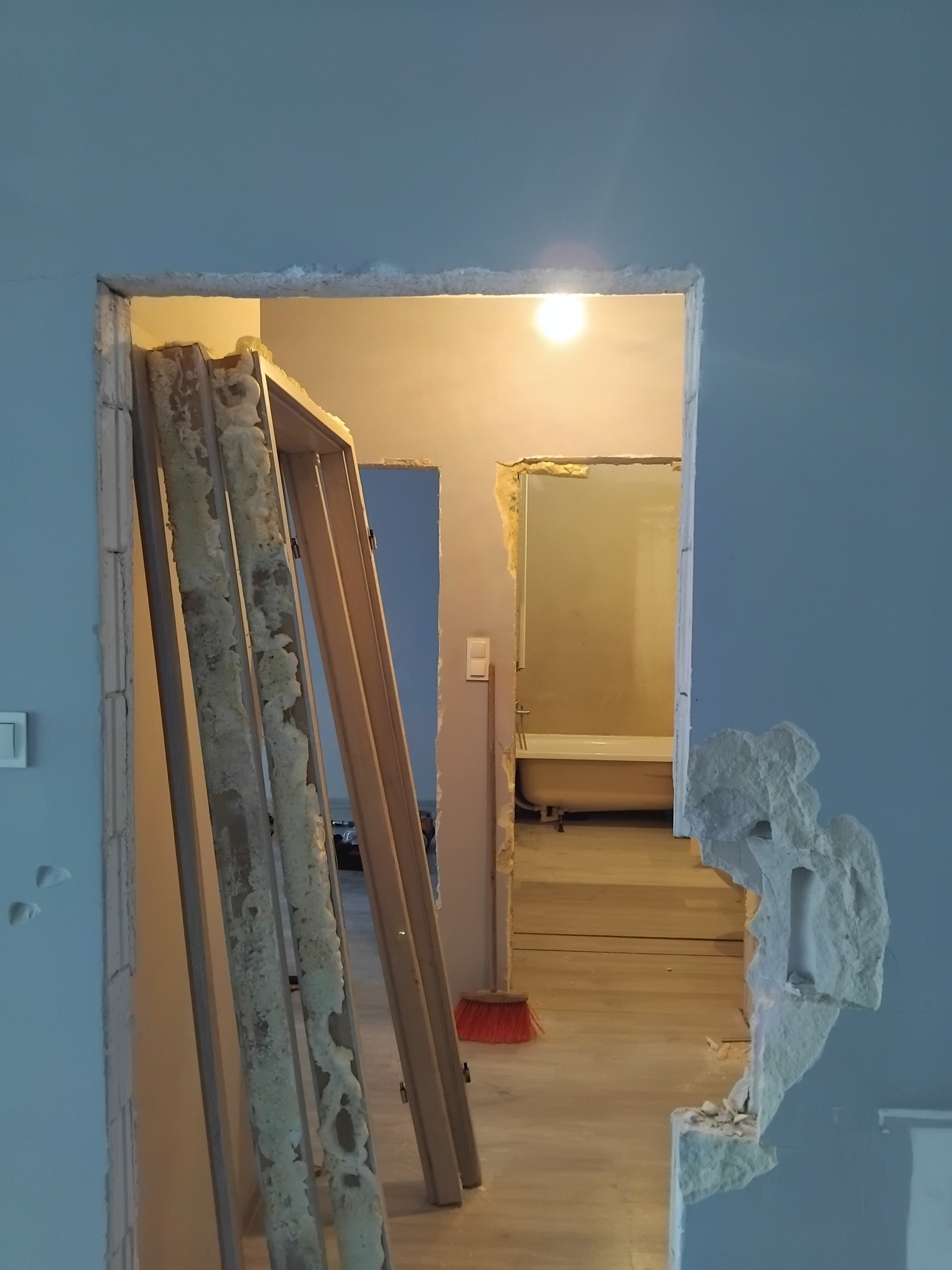 Wyburzona ściana z widoczną futryną i fragmentem łazienki w tle. Ślady po kuciu, surowy stan po demontażu. Czerwona zmiotka oparta o ścianę.