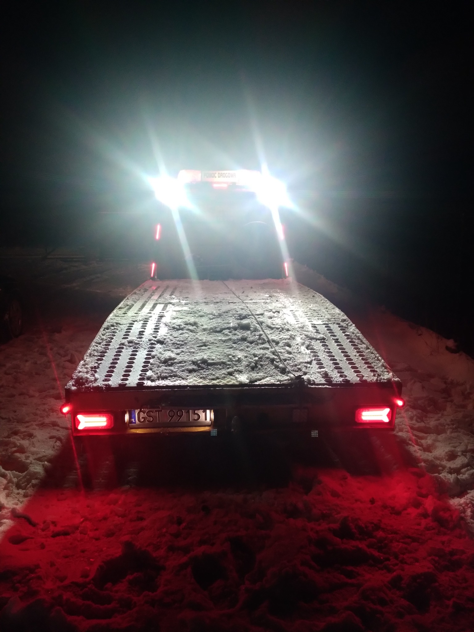 Tył lawety z tablicą rejestracyjną GST 9915I, oświetlony światłami w zimowej scenerii z padającym śniegiem.