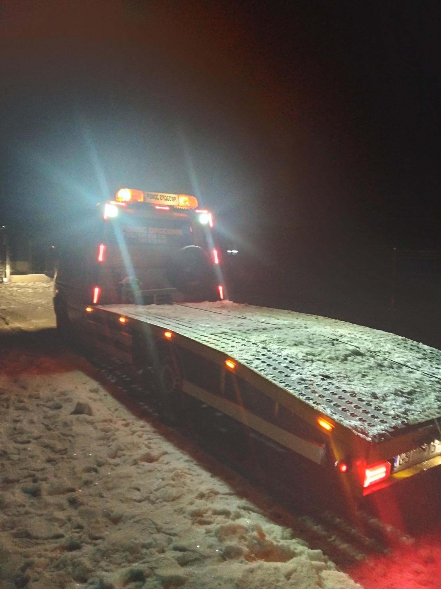 Laweta pomocy drogowej z włączonymi światłami ostrzegawczymi, zaparkowana na ośnieżonej drodze nocą.