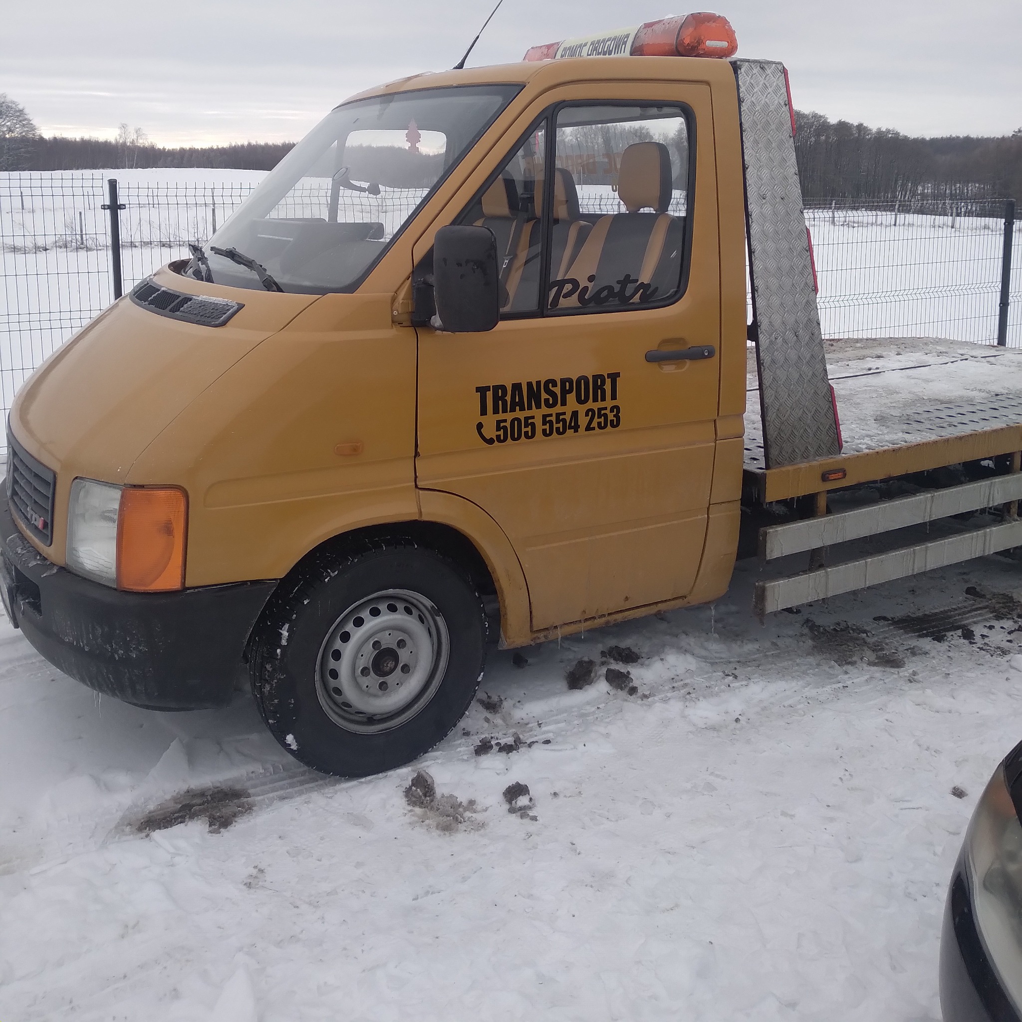 Żółty samochód dostawczy Vw lt 28 z platformą lawetową, zaparkowany na śniegu, z napisem 'TRANSPORT' i numerem telefonu na drzwiach kabiny.