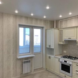 Serhii Melnyk - Jasna kuchnia z kremowymi szafkami, płytą gazową, piekarnikiem i oknem balkonowym z grzejnikiem pod spodem. Ściany pokryte tapetą w delikatny wzór, sufit z wbudowanymi punktowymi lampami.