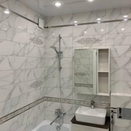 Serhii Melnyk - Wykończona łazienka z białymi płytkami imitującymi marmur, ozdobnymi bordiurami, baterią wannową, umywalką nablatową i szafką z lustrem, oświetlona punktowymi lampami sufitowymi.