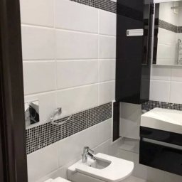 Serhii Melnyk - Nowoczesna łazienka z biało-czarnymi płytkami, wiszącą toaletą i bidetem, czarna szafka pod umywalką, widok zza brązowych drzwi.