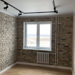 Serhii Melnyk - Pokój z dekoracyjną ścianą z cegły w odcieniach beżu i szarości, oświetlony reflektorami na szynie. Białe okno z widokiem na krajobraz. Grzejnik pod oknem i instalacja rurowa na ścianie.