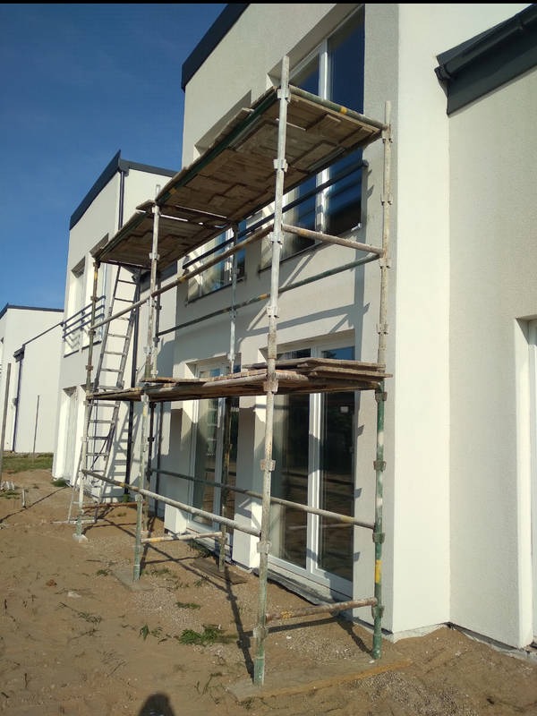 Elewacja budynku w Gdańsku podczas prac wykończeniowych, widoczne rusztowania z drewnianymi pomostami i drabina, świeżo nałożony tynk o delikatnej fakturze, okna z białymi ramami.