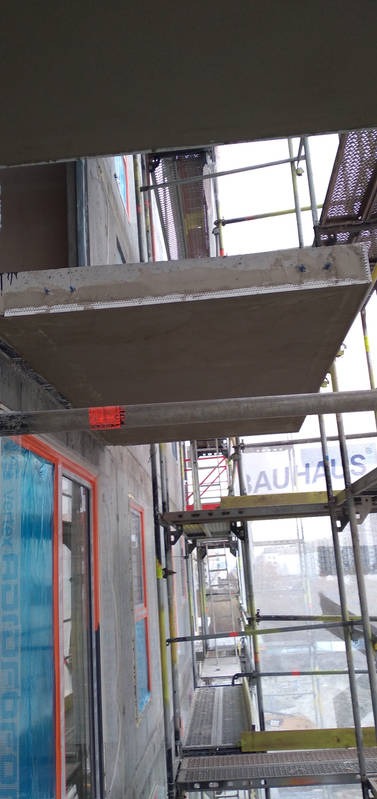 Szare elewacje budynku w trakcie ocieplania, widoczne rusztowania, okna zabezpieczone folią ochronną, fragment ocieplonego poddasza.