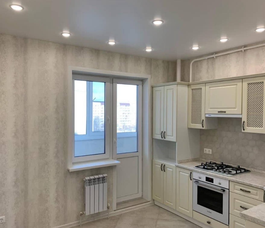 Jasna kuchnia z kremowymi szafkami, płytą gazową, piekarnikiem i oknem balkonowym z grzejnikiem pod spodem. Ściany pokryte tapetą w delikatny wzór, sufit z wbudowanymi punktowymi lampami.