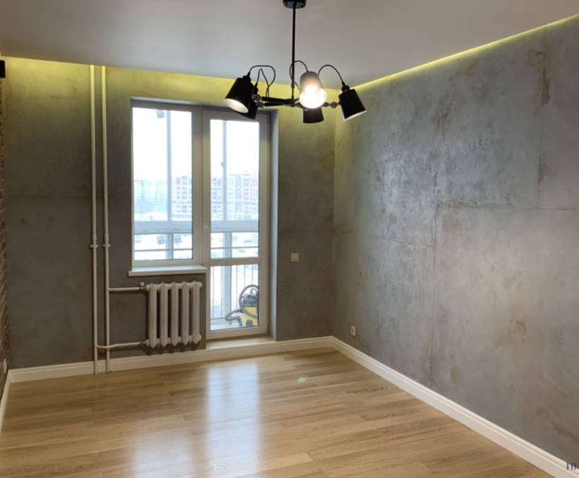 Pokój po odświeżeniu: parkiet, ściany z efektem betonu, biała listwa przypodłogowa, okno z grzejnikiem, designerska lampa sufitowa.
