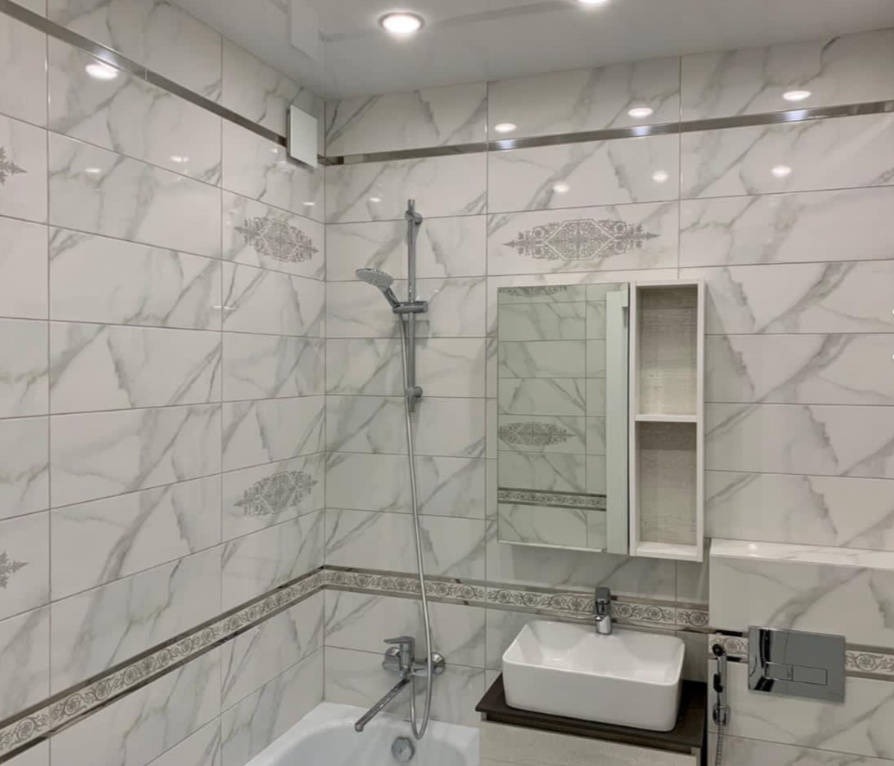 Wykończona łazienka z białymi płytkami imitującymi marmur, ozdobnymi bordiurami, baterią wannową, umywalką nablatową i szafką z lustrem, oświetlona punktowymi lampami sufitowymi.