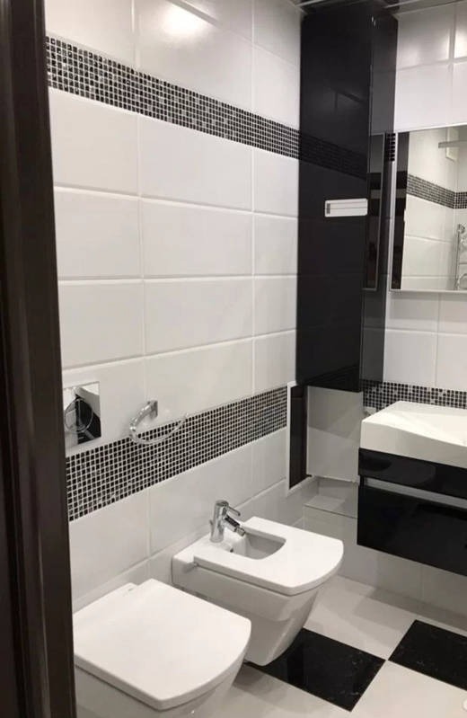 Nowoczesna łazienka z biało-czarnymi płytkami, wiszącą toaletą i bidetem, czarna szafka pod umywalką, widok zza brązowych drzwi.