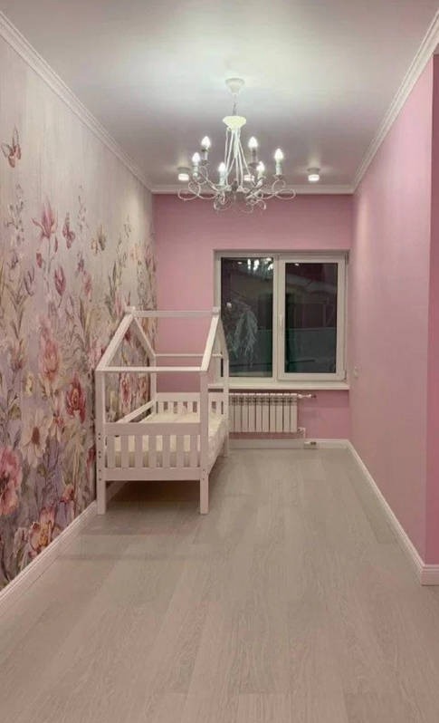 Pokój dziecięcy z łóżkiem w kształcie domku, różowymi ścianami i tapetą w kwiaty, oświetlony żyrandolem z białymi abażurami.