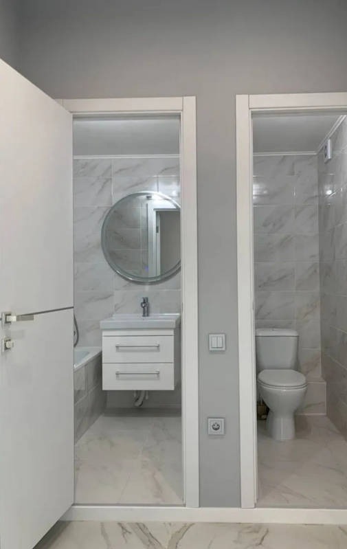 Dwa wejścia do łazienki wykończonej białymi płytkami imitującymi marmur, po lewej stronie umywalka z okrągłym lustrem, po prawej toaleta.