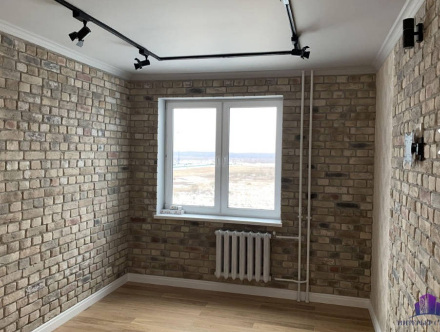 Pokój z dekoracyjną ścianą z cegły w odcieniach beżu i szarości, oświetlony reflektorami na szynie. Białe okno z widokiem na krajobraz. Grzejnik pod oknem i instalacja rurowa na ścianie.