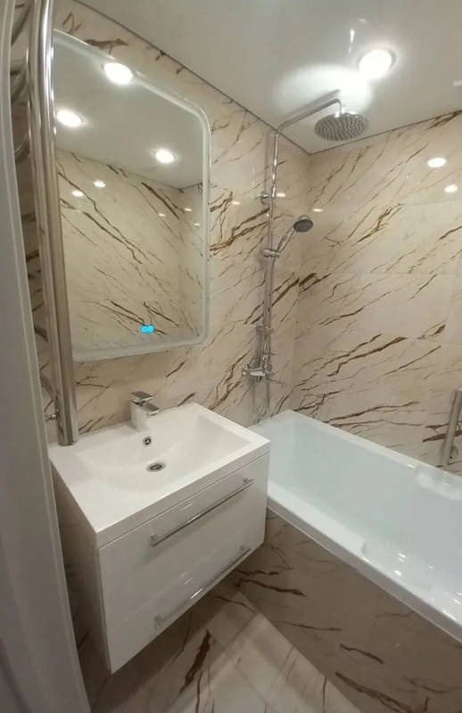 Wykończona łazienka z marmurowymi płytkami, białą wanną, umywalką z szafką i lustrem z podświetleniem LED.