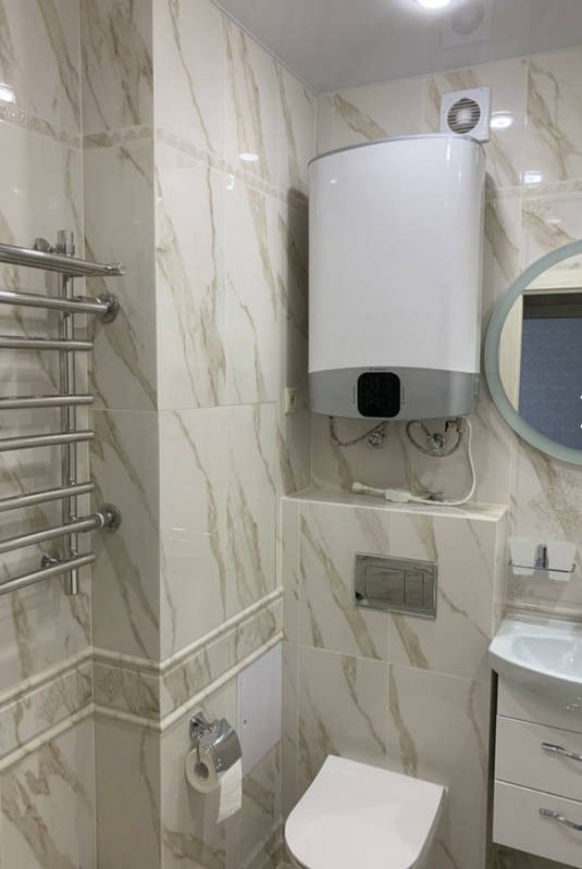 Wykończona łazienka z marmurowymi płytkami, podgrzewanym wieszakiem na ręczniki, białą umywalką z szafką i bojlerem Ariston zamontowanym na ścianie.