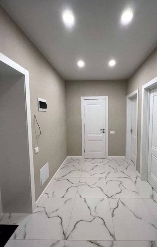 Wyremontowany korytarz z białymi płytkami imitującymi marmur, kremowe ściany, białe drzwi i wpuszczane oświetlenie sufitowe.