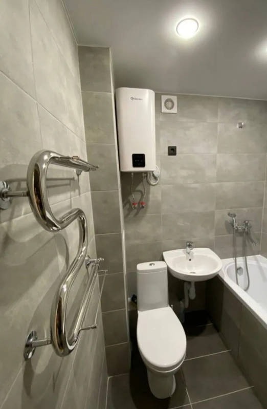 Wyremontowana łazienka z szarymi płytkami, chromowanym grzejnikiem drabinkowym, białym bojlerem elektrycznym, umywalką i toaletą.