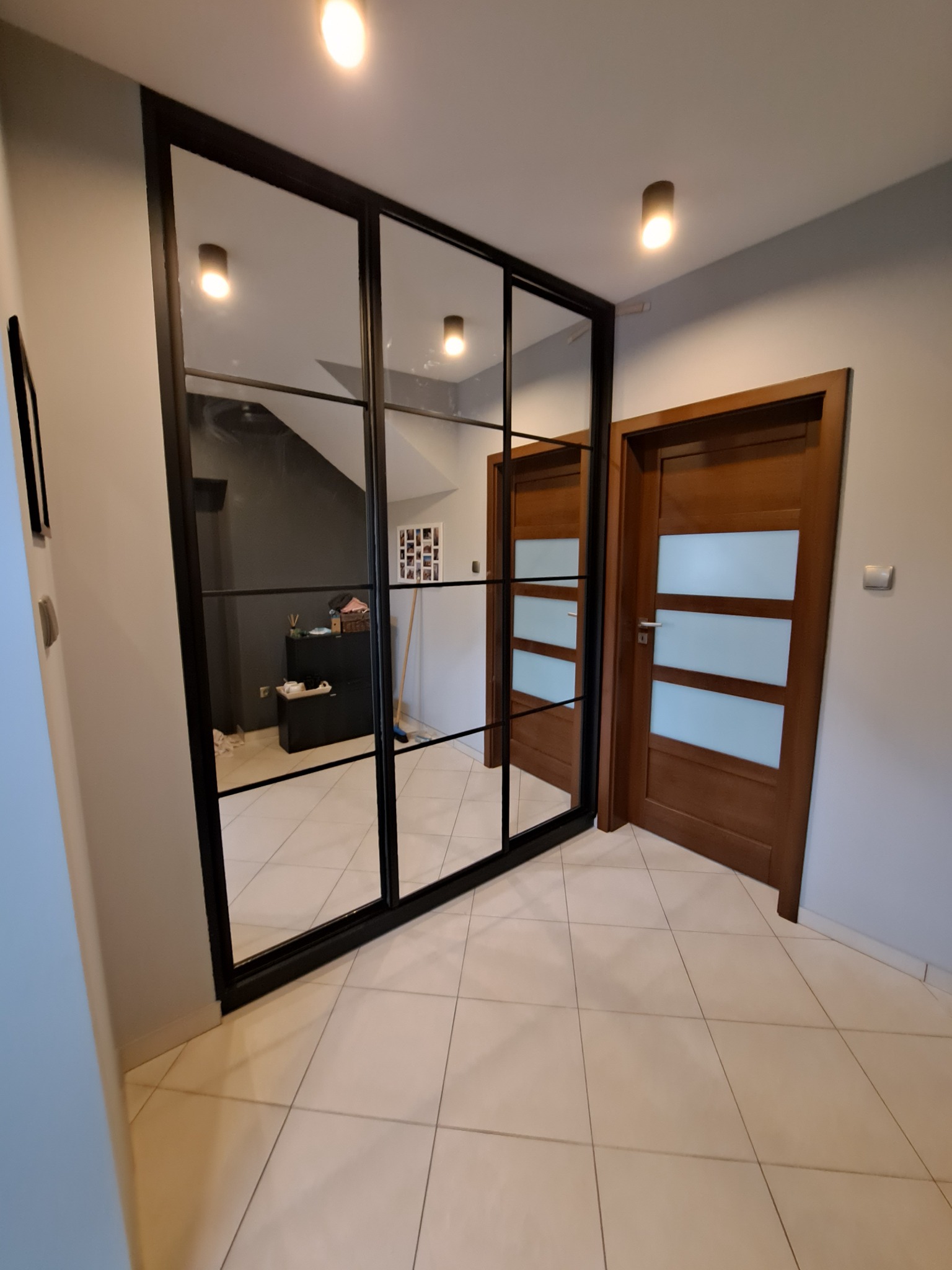 Szafa wnękowa z czarnymi ramami i lustrzanymi drzwiami przesuwnymi, odbijająca wnętrze domu, widoczna obok drewnianych drzwi z szybami, na tle jasnych płytek podłogowych.