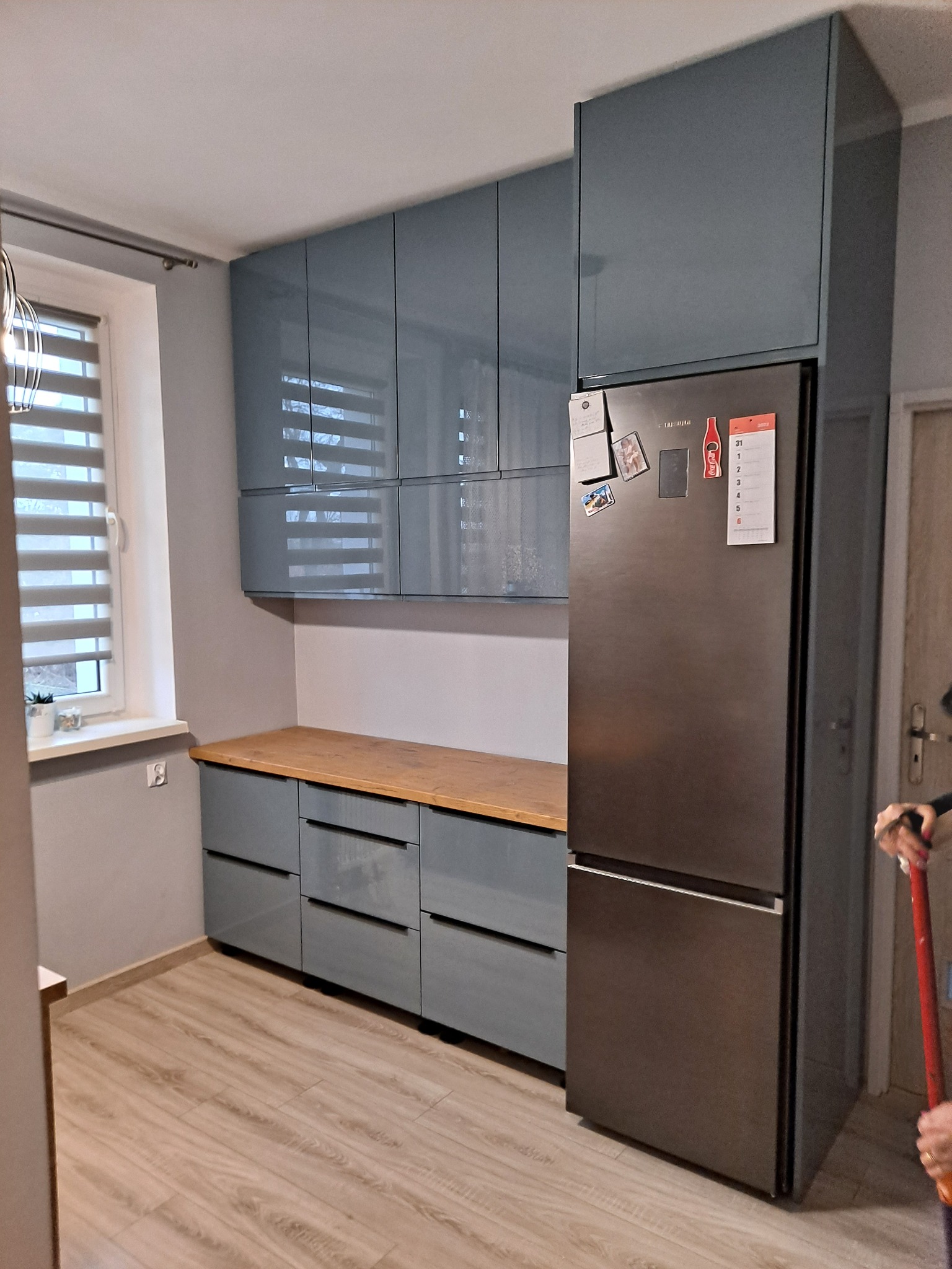 Szare, lakierowane szafki kuchenne z drewnianym blatem, połączone z wysoką zabudową lodówki, widok z perspektywy podłogi, w tle okno z żaluzjami.