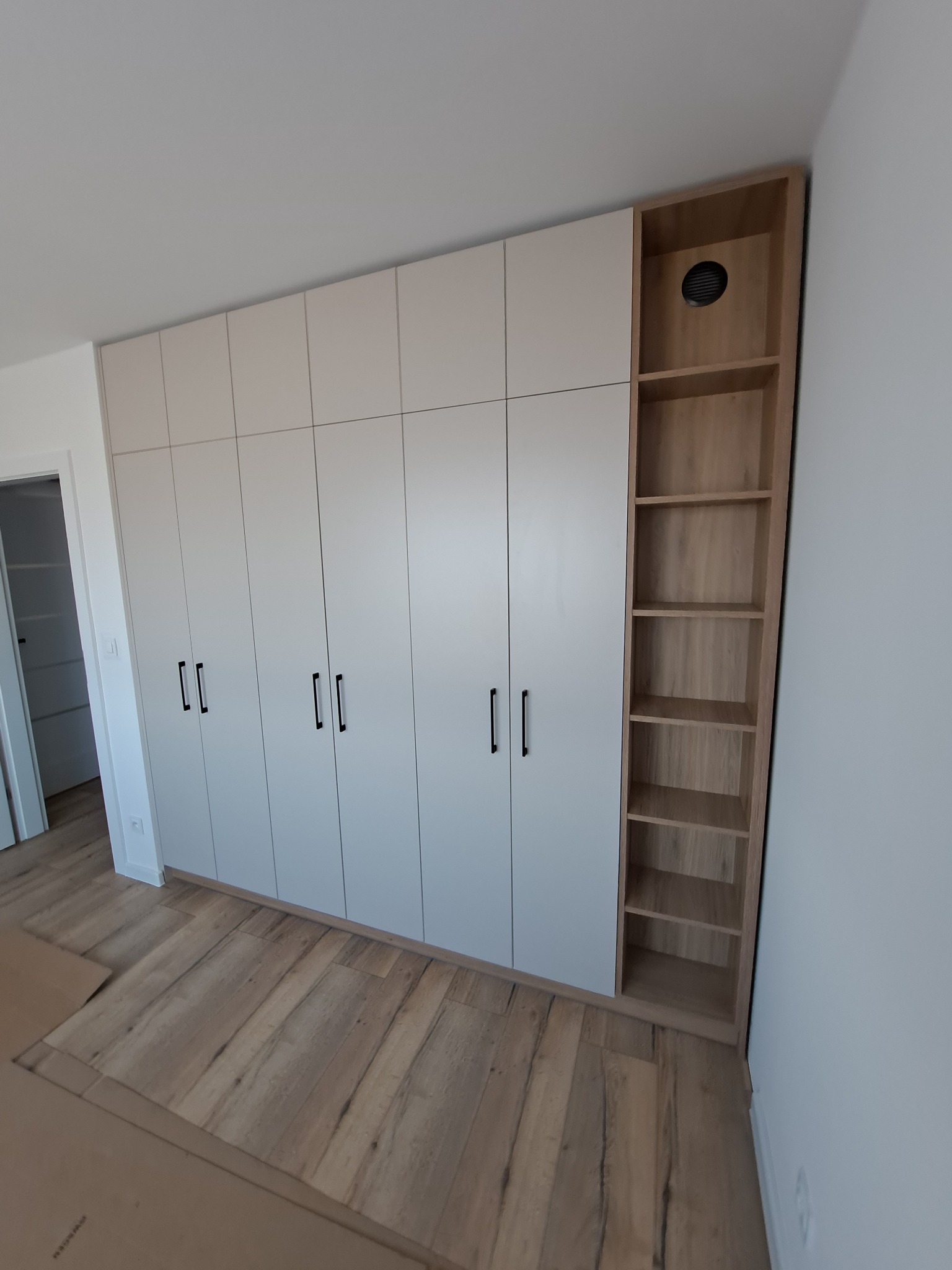 Biała szafa na wymiar z czarnymi uchwytami i otwartą, drewnianą półką z kratką wentylacyjną u góry, na drewnianej podłodze.