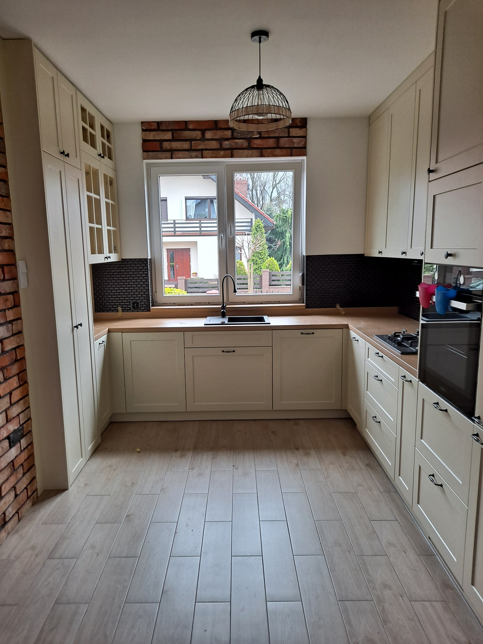 Jasna kuchnia z kremowymi szafkami, drewnianym blatem i ceglaną ścianą, widok na okno z czarną armaturą i lampą wiszącą.
