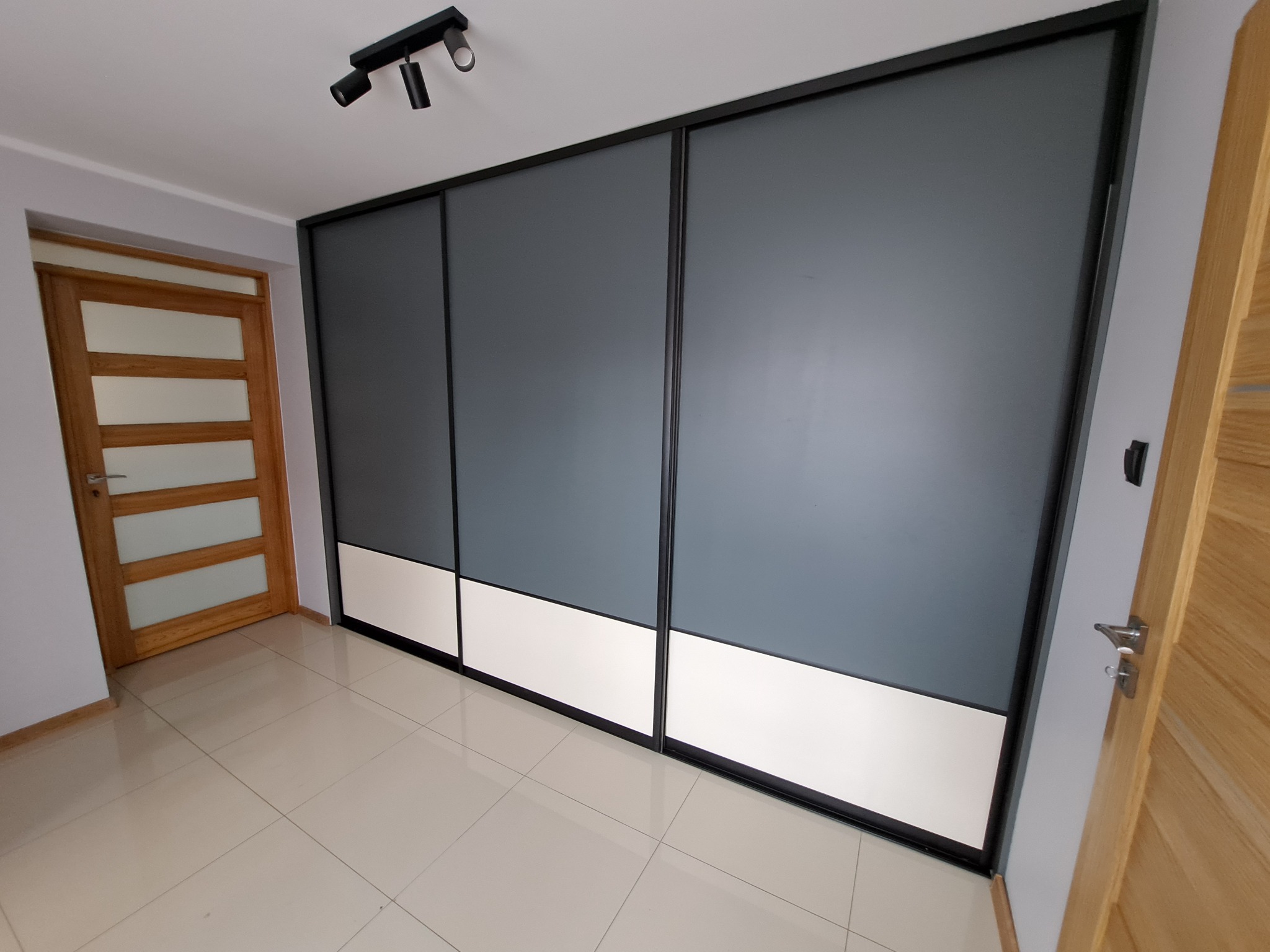 Trzydrzwiowa szafa przesuwna z szarymi frontami i białym pasem u dołu, czarne obramowanie, na jasnych płytkach podłogowych, widoczna lampa sufitowa i fragment drewnianych drzwi.