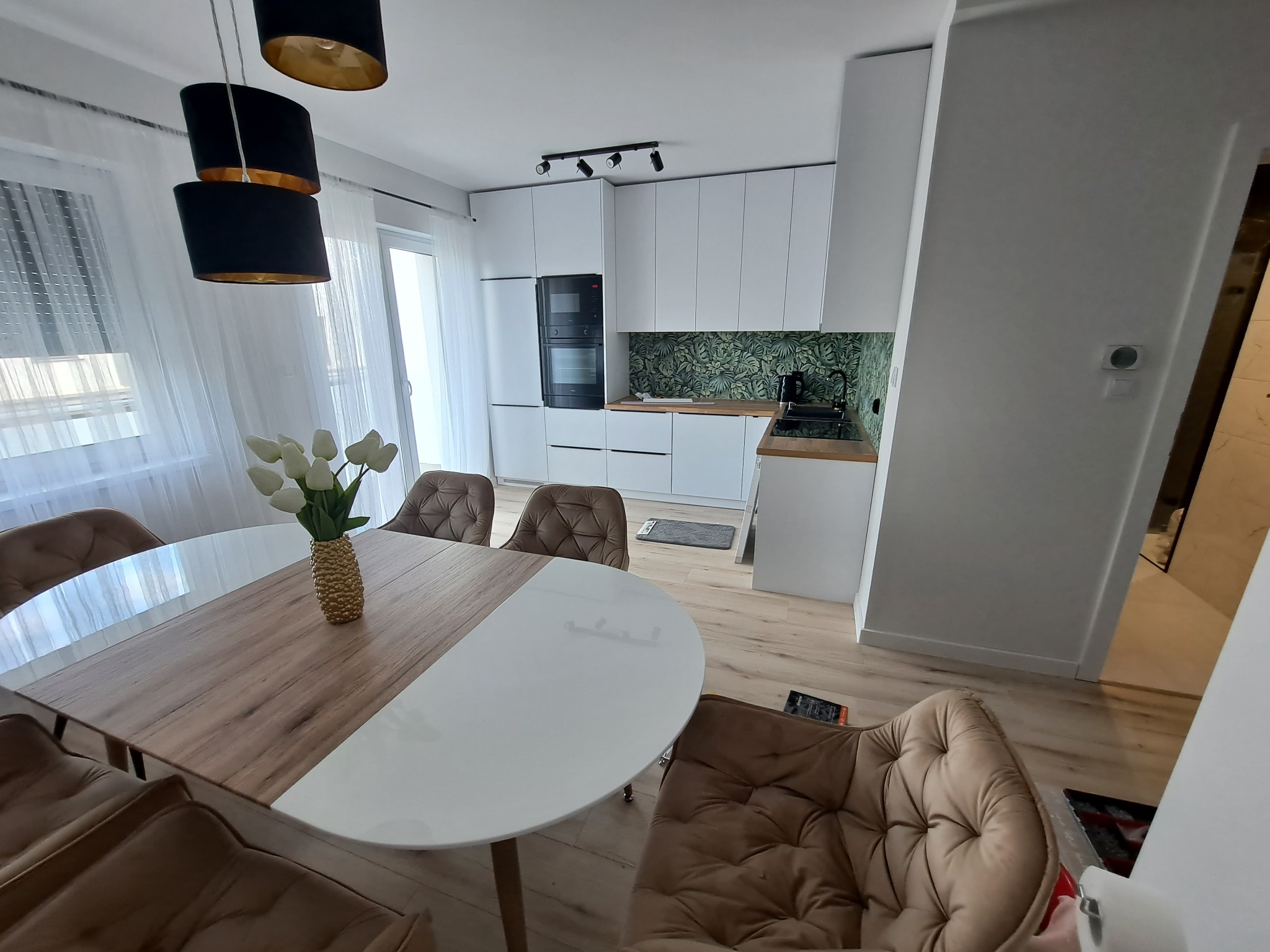 Nowoczesna kuchnia z białymi szafkami, zielonym motywem roślinnym na ścianie nad blatem, czarnym piekarnikiem i okrągłym stołem z drewnianym i białym blatem oraz brązowymi krzesłami w stylu glamour.