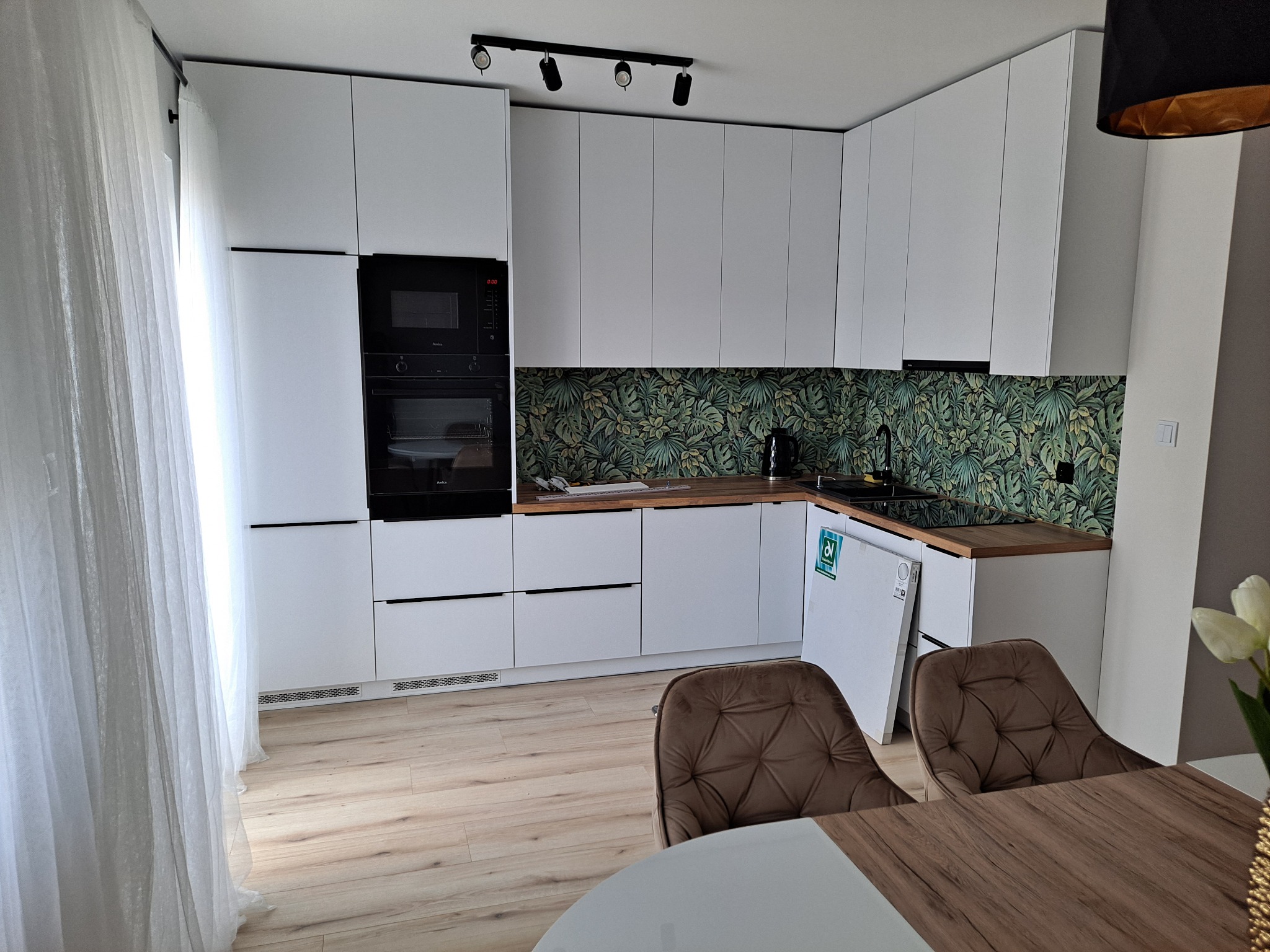 Nowoczesna kuchnia z białymi, matowymi frontami, czarnym piekarnikiem i płytą indukcyjną, blatem z jasnego drewna i zieloną tapetą w tropikalne liście, oświetlona reflektorami sufitowymi i światłem...