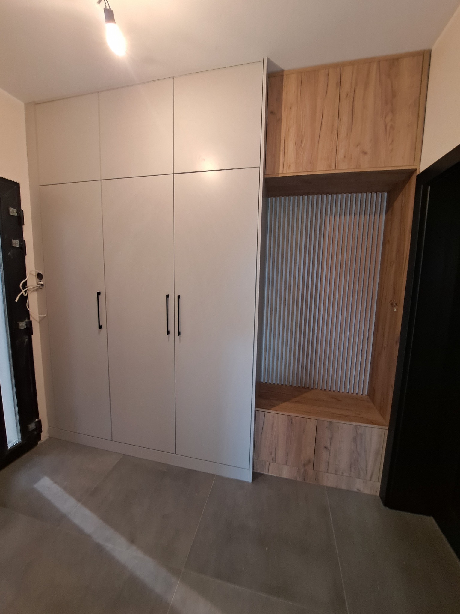 Nowoczesna szafa w przedpokoju z białymi frontami i drewnianym akcentem w postaci siedziska z pionowymi listwami dekoracyjnymi.