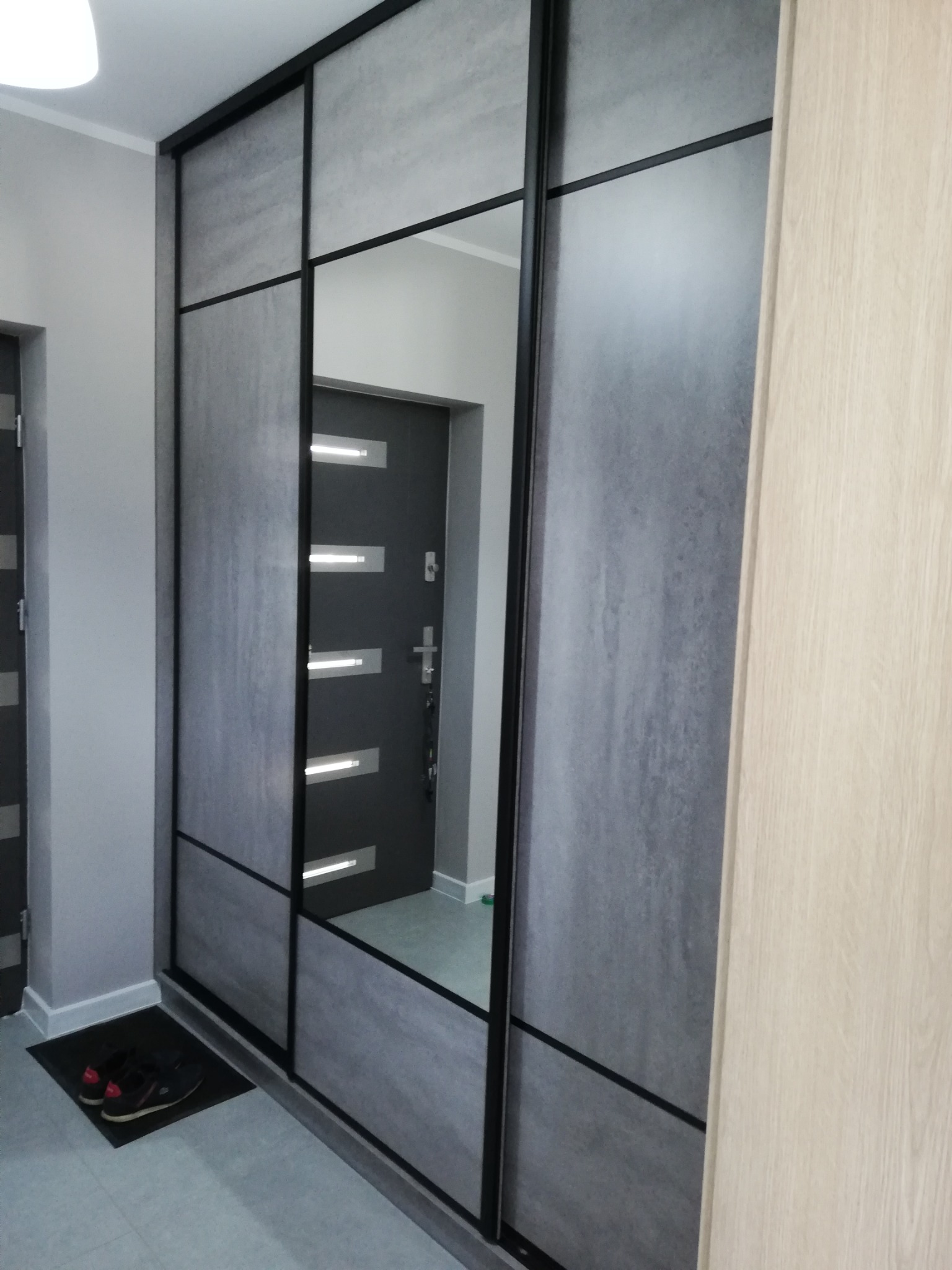 Szafa wnękowa z przesuwnymi drzwiami o szarym, betonowym wykończeniu i czarnych profilach, z lustrem odbijającym drzwi wejściowe do mieszkania. Na podłodze leżą czarne buty na wycieraczce.