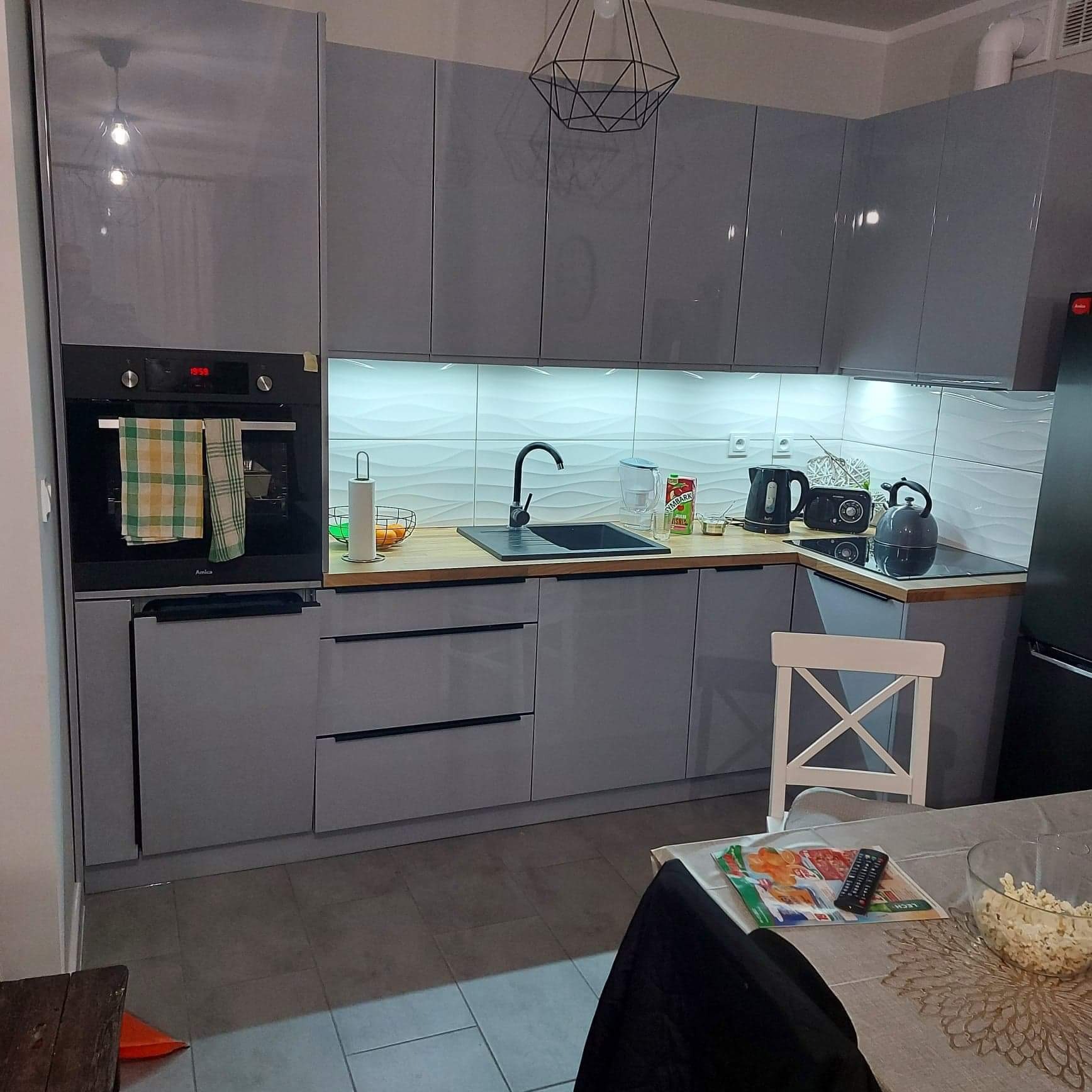 Szara, nowoczesna kuchnia z błyszczącymi frontami, czarnym zlewem, drewnianym blatem i białymi płytkami z falistym wzorem na ścianie, oświetlona pod szafkami.