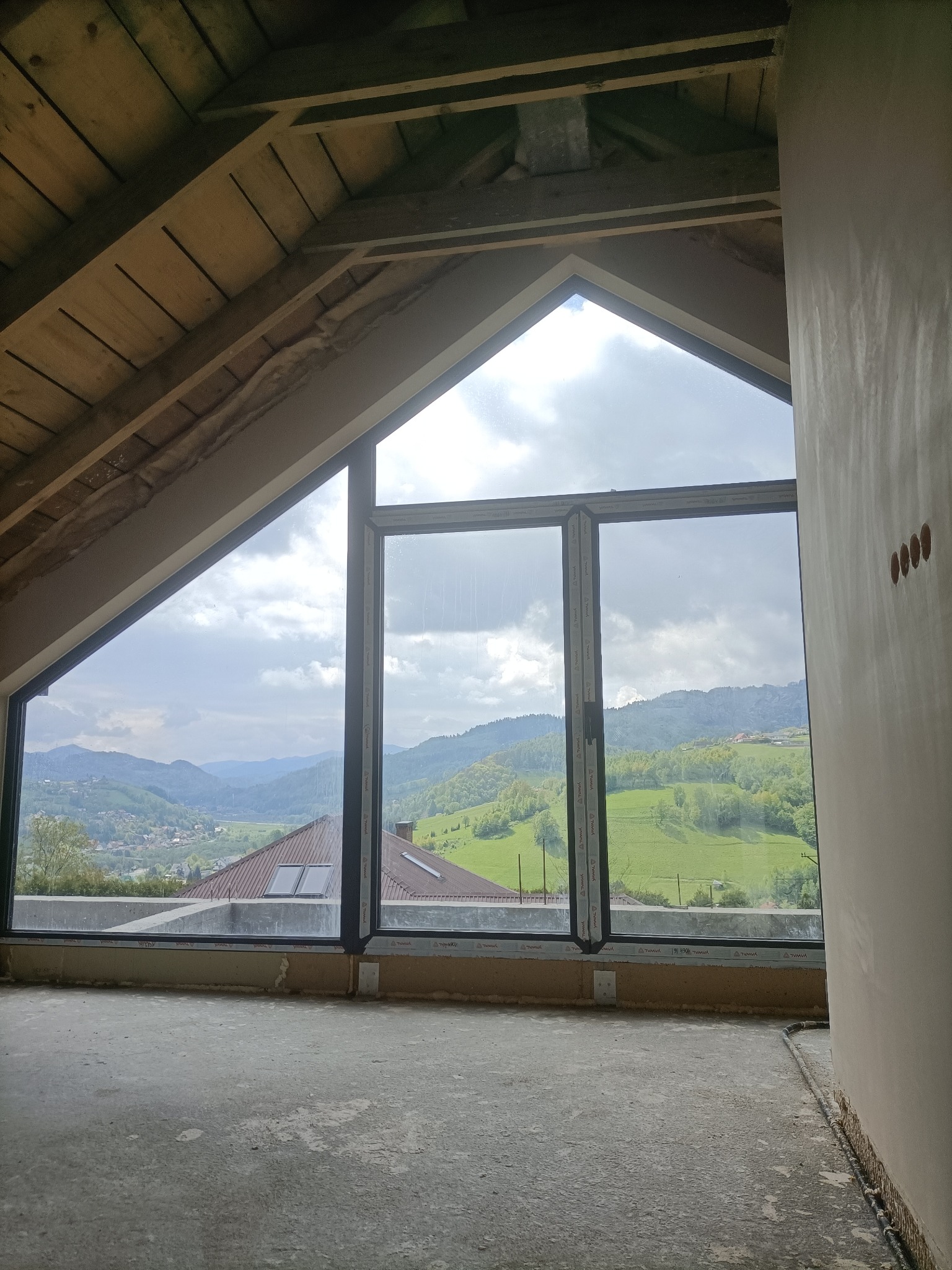 Wnętrze poddasza z dużym trójkątnym oknem z widokiem na górzysty krajobraz, surowy stan wykończenia ścian i podłogi, widoczne drewniane elementy konstrukcji dachu.