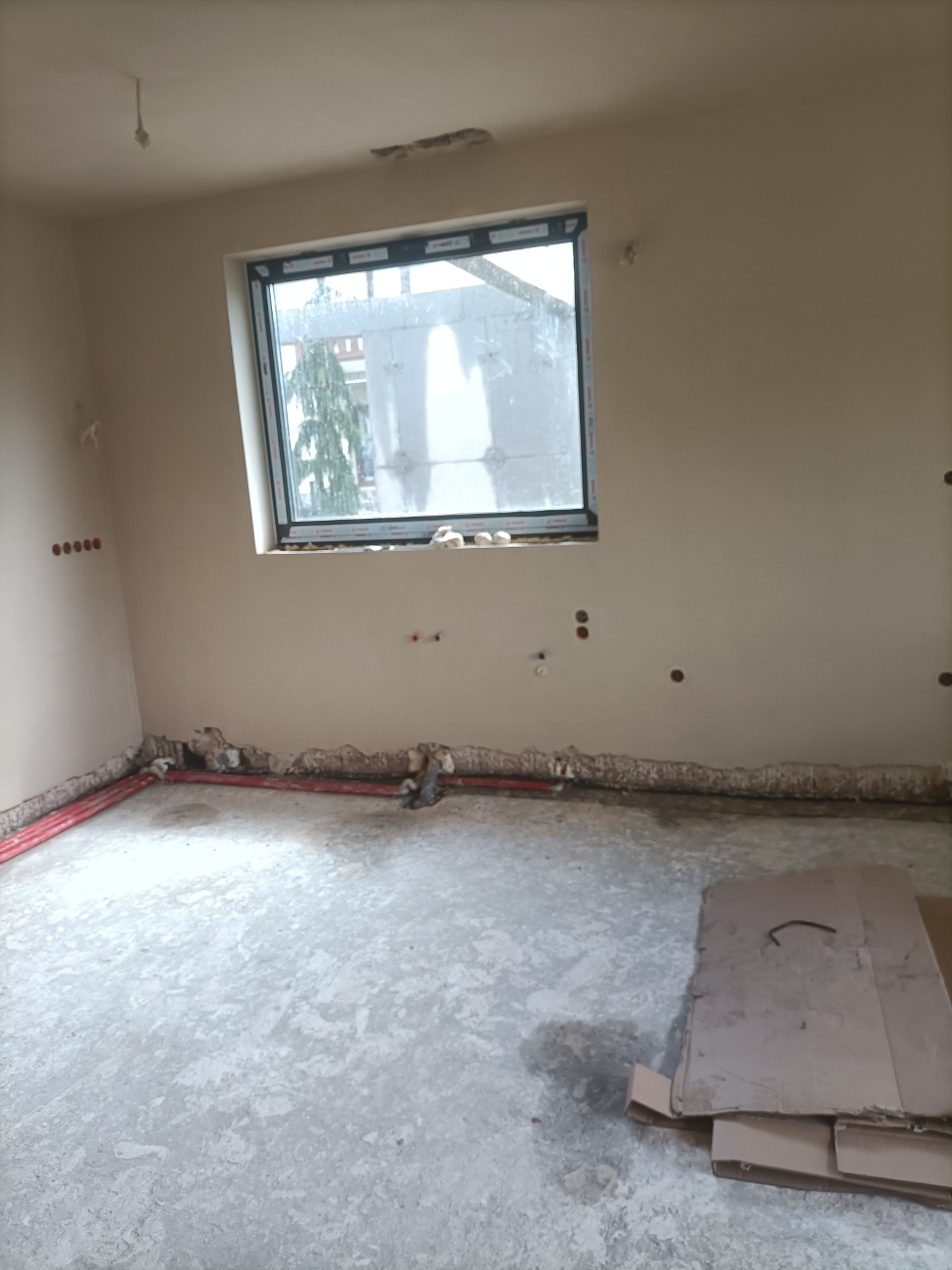 Pokój w stanie surowym: ściany przygotowane pod malowanie, widoczne instalacje elektryczne i hydrauliczne przy podłodze, okno z widokiem na zewnątrz, podłoga betonowa z resztkami materiałów...