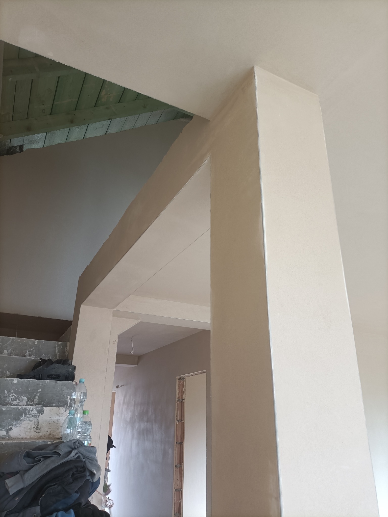 Wnętrze w trakcie wykańczania: obłożona płytami gipsowymi kolumna i fragment sufitu, widoczne schody z butelkami wody i ubraniami, ściany przygotowane do malowania.