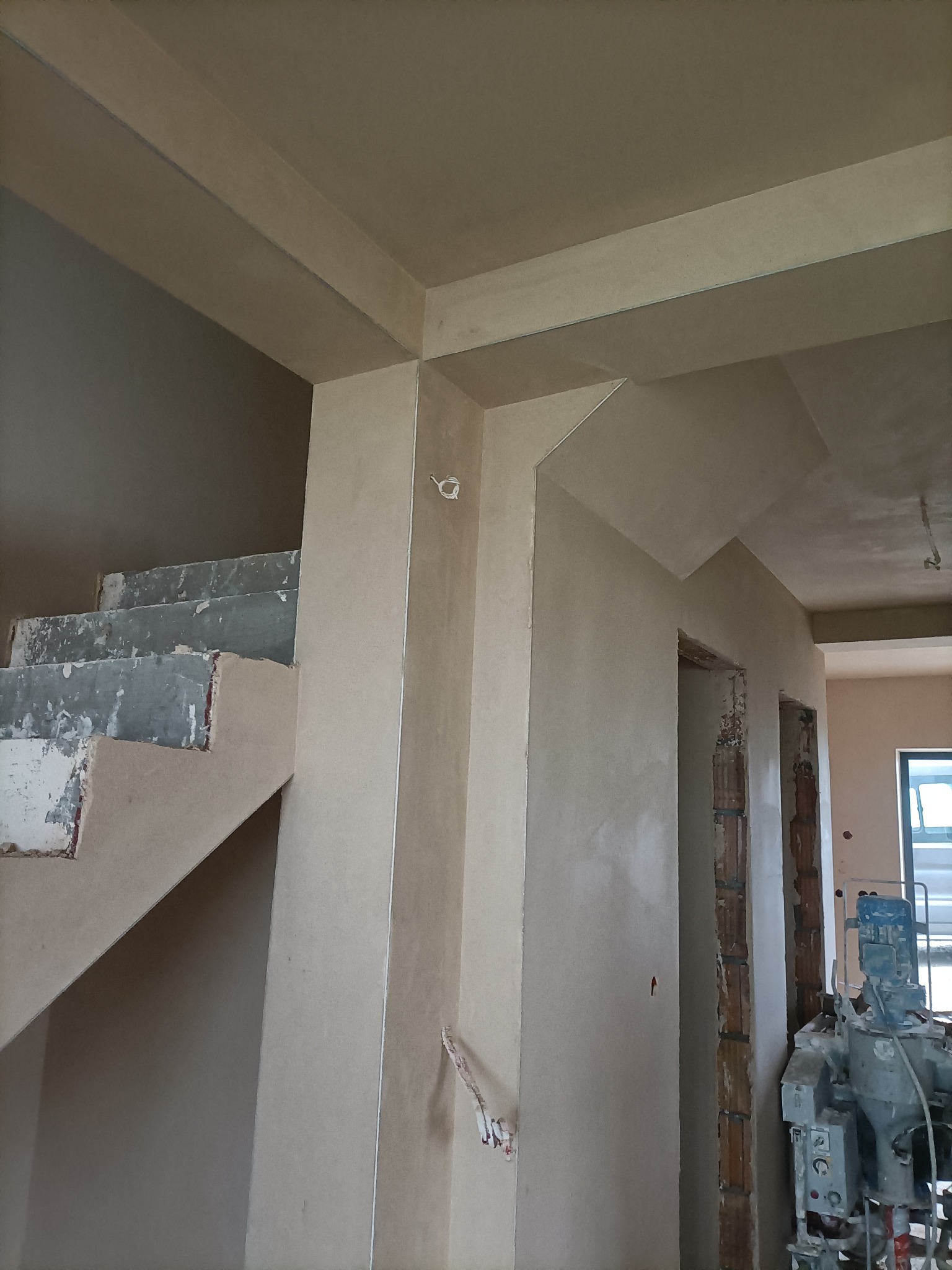 Wnętrze w trakcie tynkowania maszynowego, widoczne surowe schody, świeżo otynkowane ściany i fragment maszyny do tynkowania.