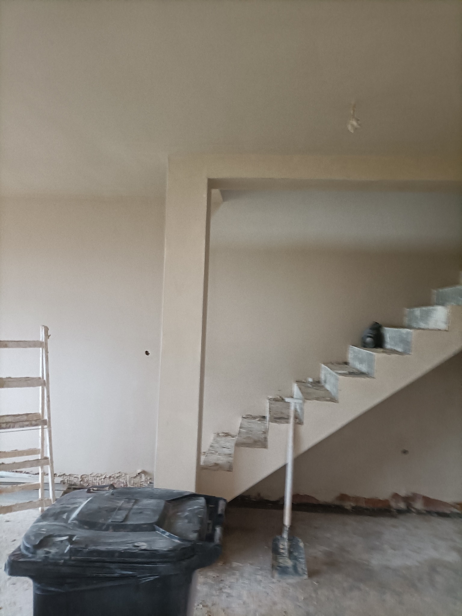 Wnętrze w trakcie prac wykończeniowych: ściany po nałożeniu tynku, widoczne schody w surowym stanie, drabina oparta o ścianę oraz czarny pojemnik na odpady budowlane na pierwszym planie.