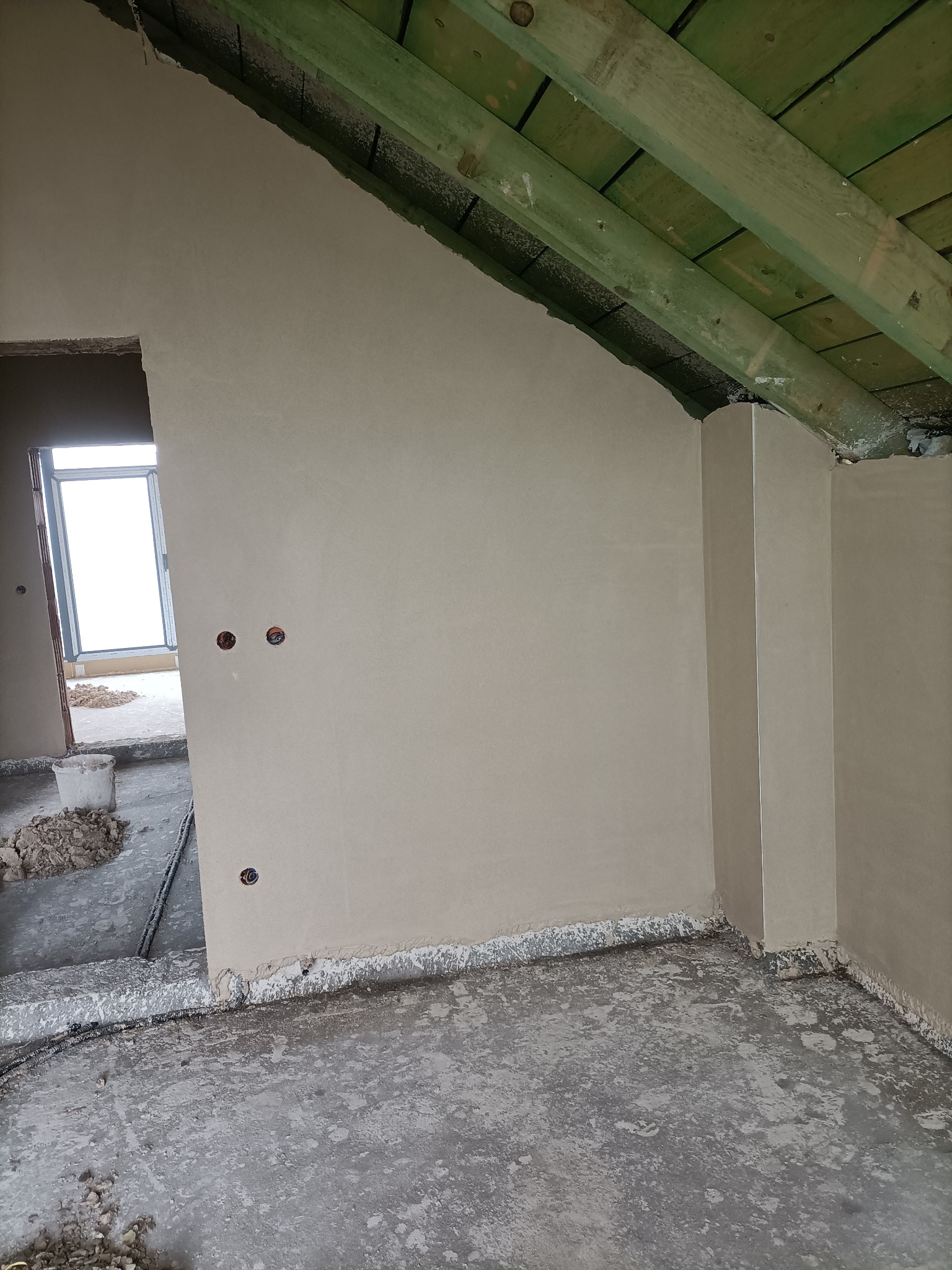 Świeżo nałożona, gładka warstwa tynku na ścianach poddasza z widocznymi puszkami elektrycznymi i surową, drewnianą konstrukcją dachu. Podłoże betonowe z resztkami materiałów budowlanych.