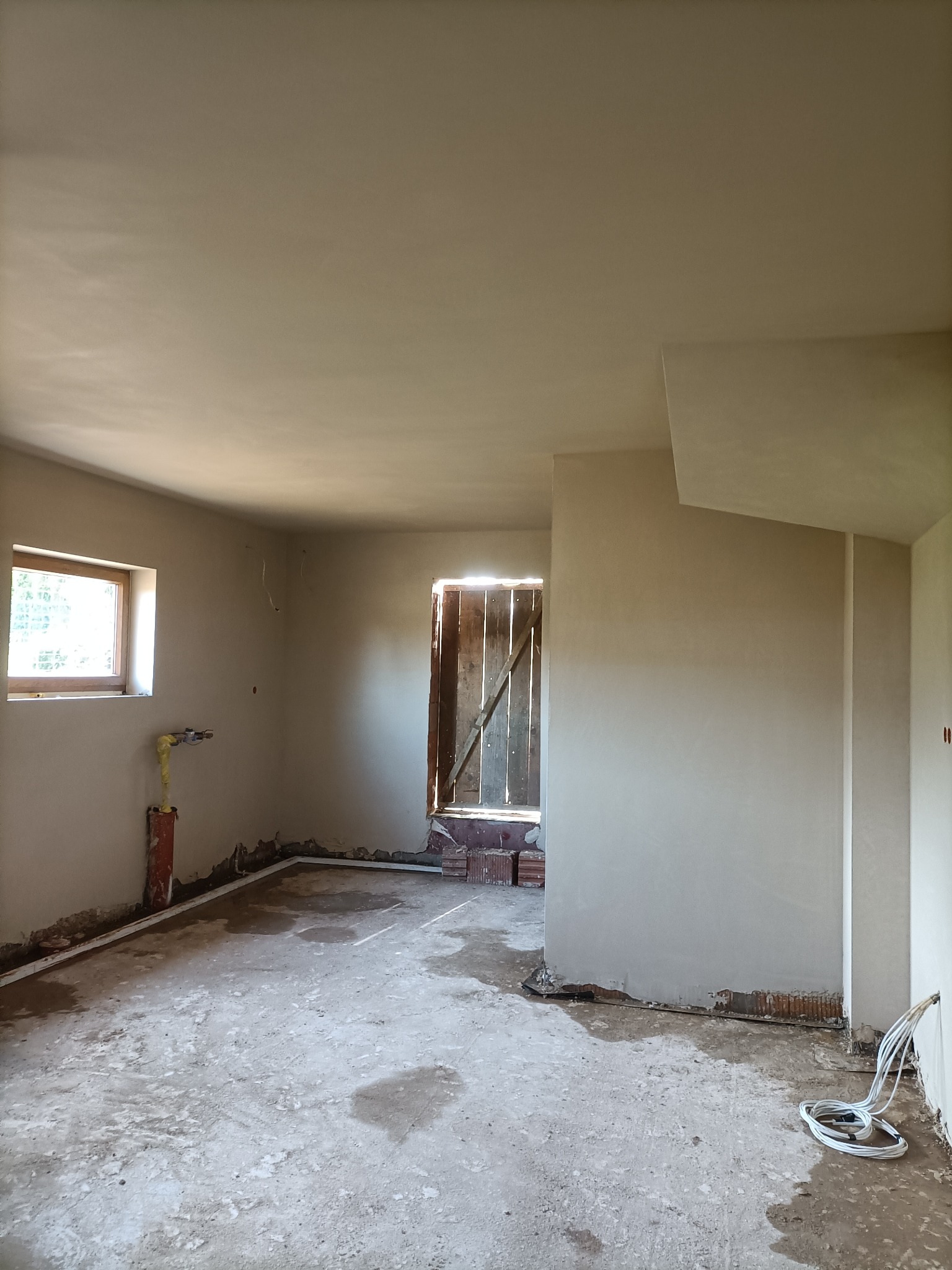 Wnętrze w trakcie prac wykończeniowych: ściany pokryte świeżym tynkiem, widoczne instalacje, fragmenty cegieł i przewody na podłodze, okno z drewnianą ramą i drzwi z desek w tle.