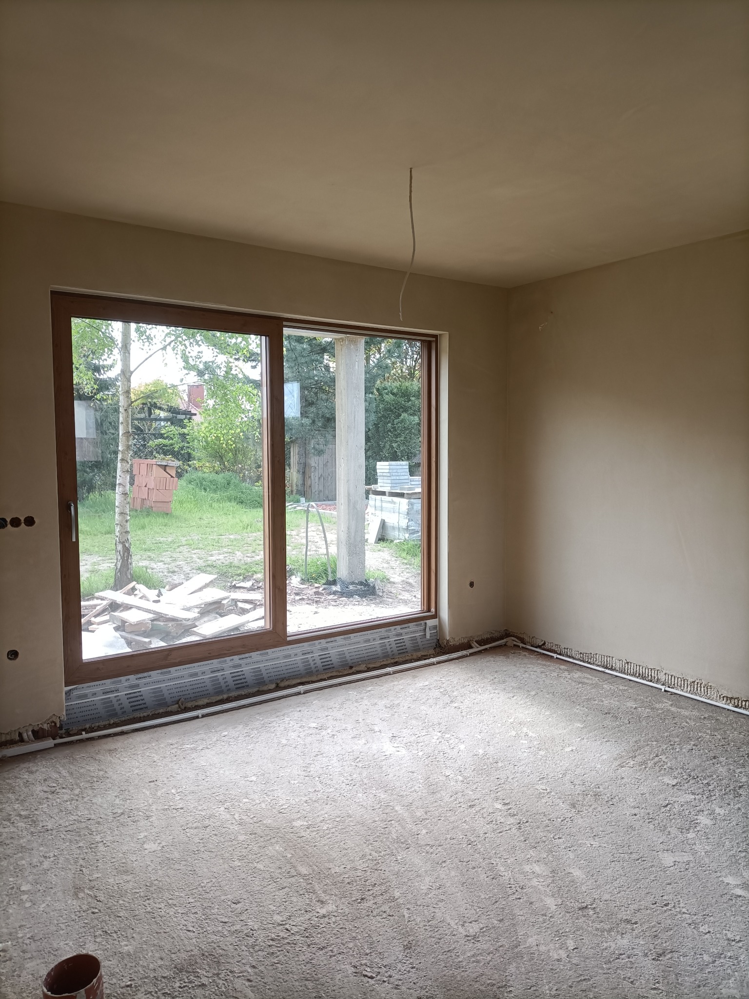 Wnętrze w stanie deweloperskim z oknem przesuwnym, ściany pokryte tynkiem, widoczne instalacje podłogowe i fragment wiaderka w rogu.