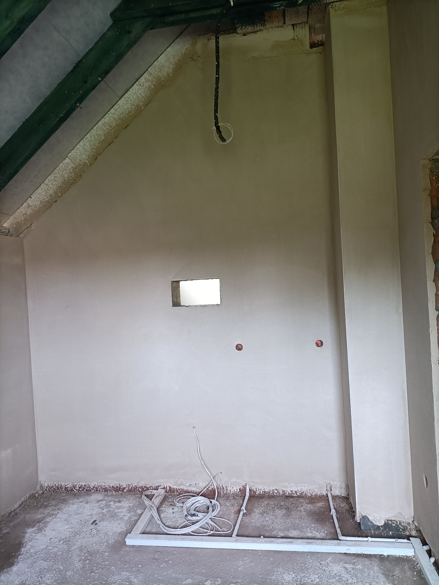 Wnętrze poddasza po nałożeniu jasnej warstwy gładzi, widoczne wystające kable elektryczne, mały otwór okienny i surowe drewniane elementy konstrukcyjne.