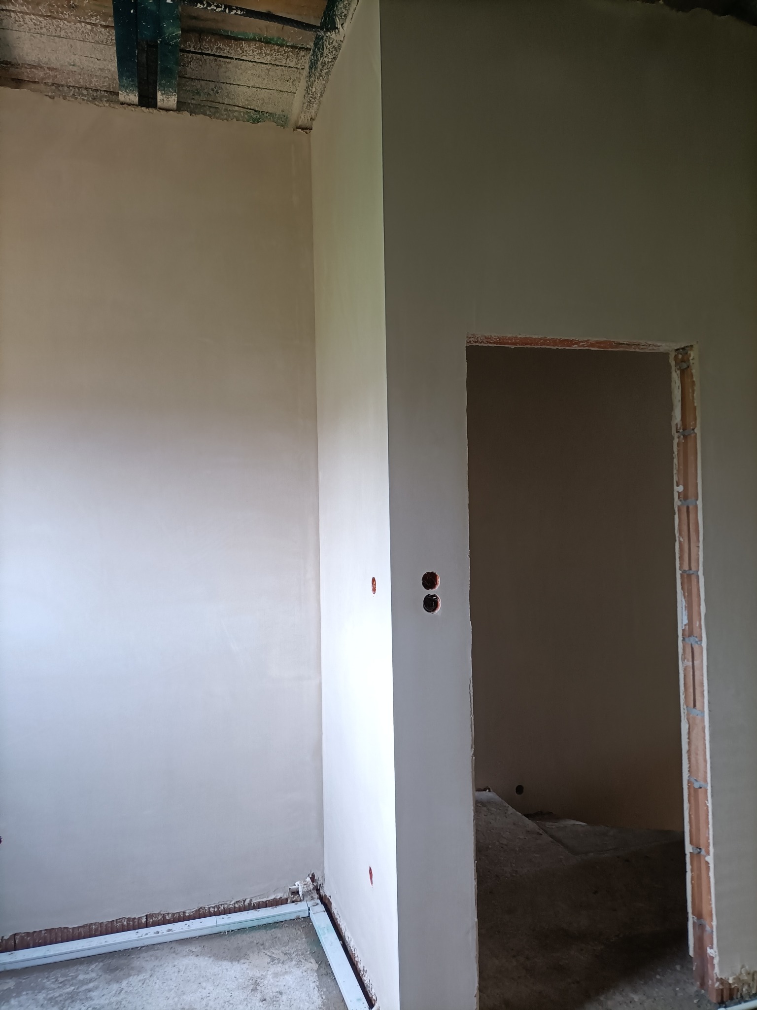 Wnętrze w trakcie prac wykończeniowych: ściany pokryte świeżo nałożoną, jasną warstwą, widoczne puszki elektryczne i ramy ościeżnic przed montażem drzwi, surowy sufit z drewnianymi belkami.