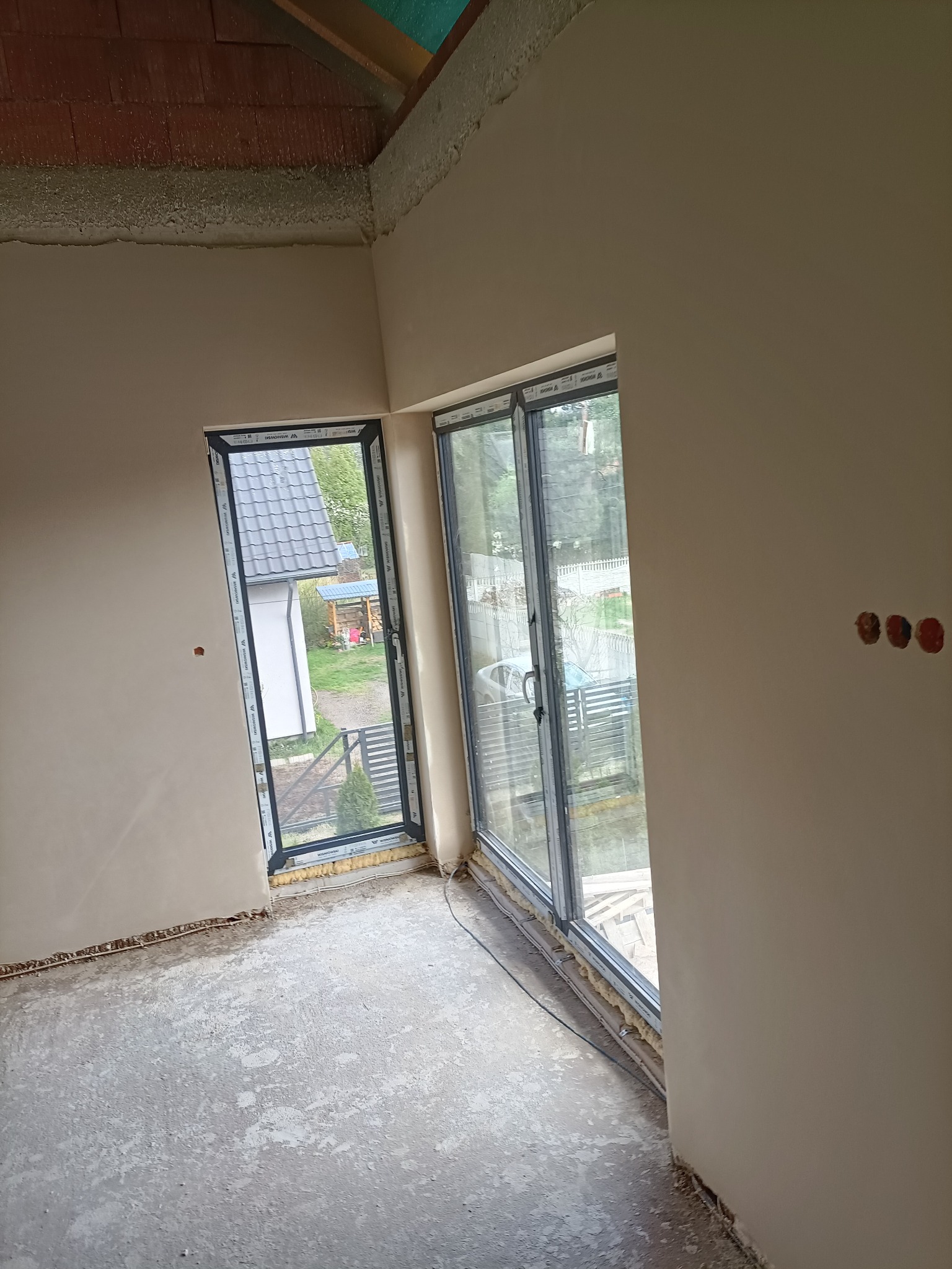 Wnętrze domu w trakcie prac wykończeniowych: ściany pokryte gładzią, okna osadzone, surowa podłoga przed wylewką, widoczne puszki elektryczne.