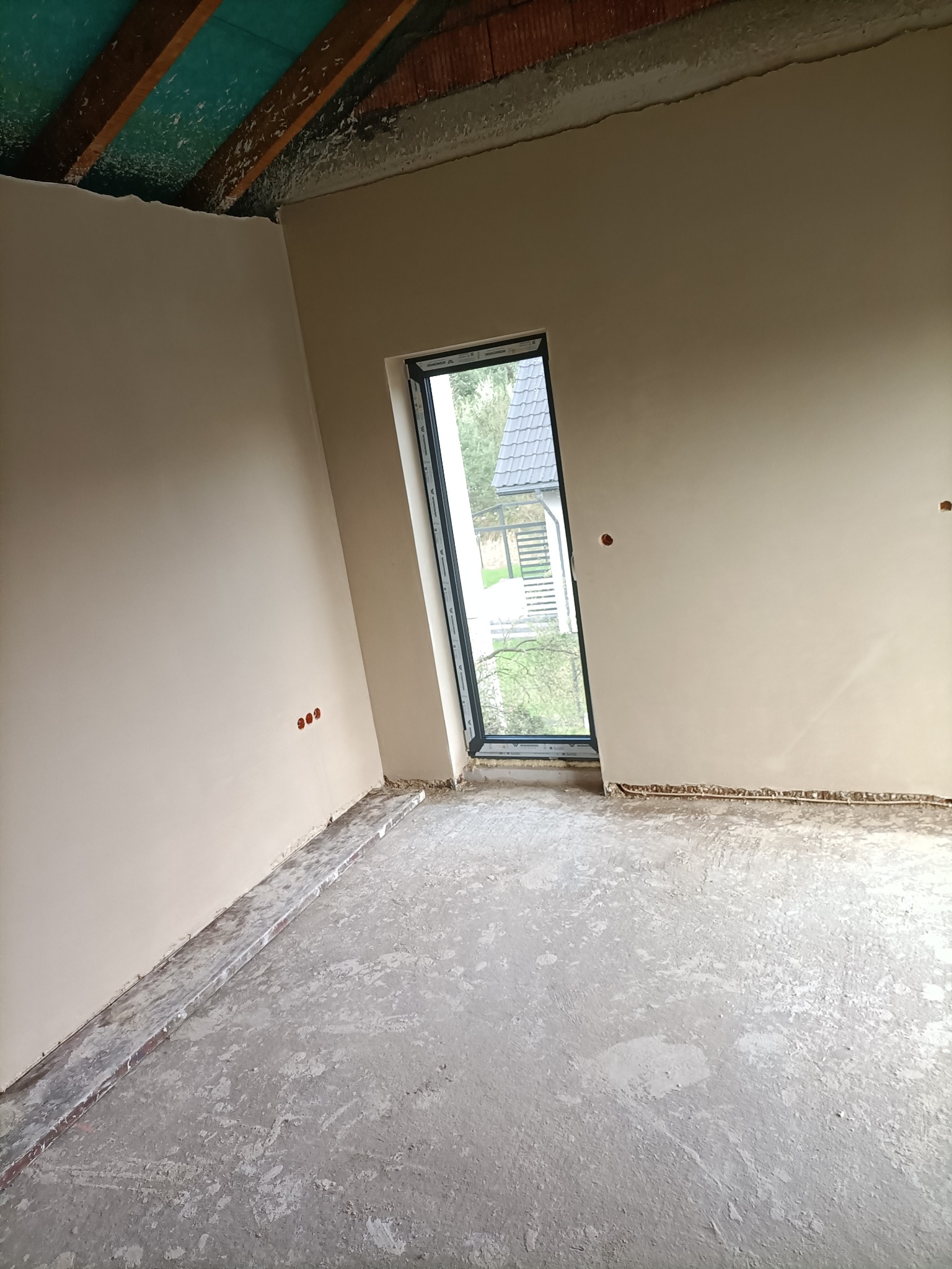 Wnętrze w stanie deweloperskim z gładkimi, jasnymi ścianami po szpachlowaniu, surowy beton na podłodze, okno z widokiem na dom z szarą dachówką.