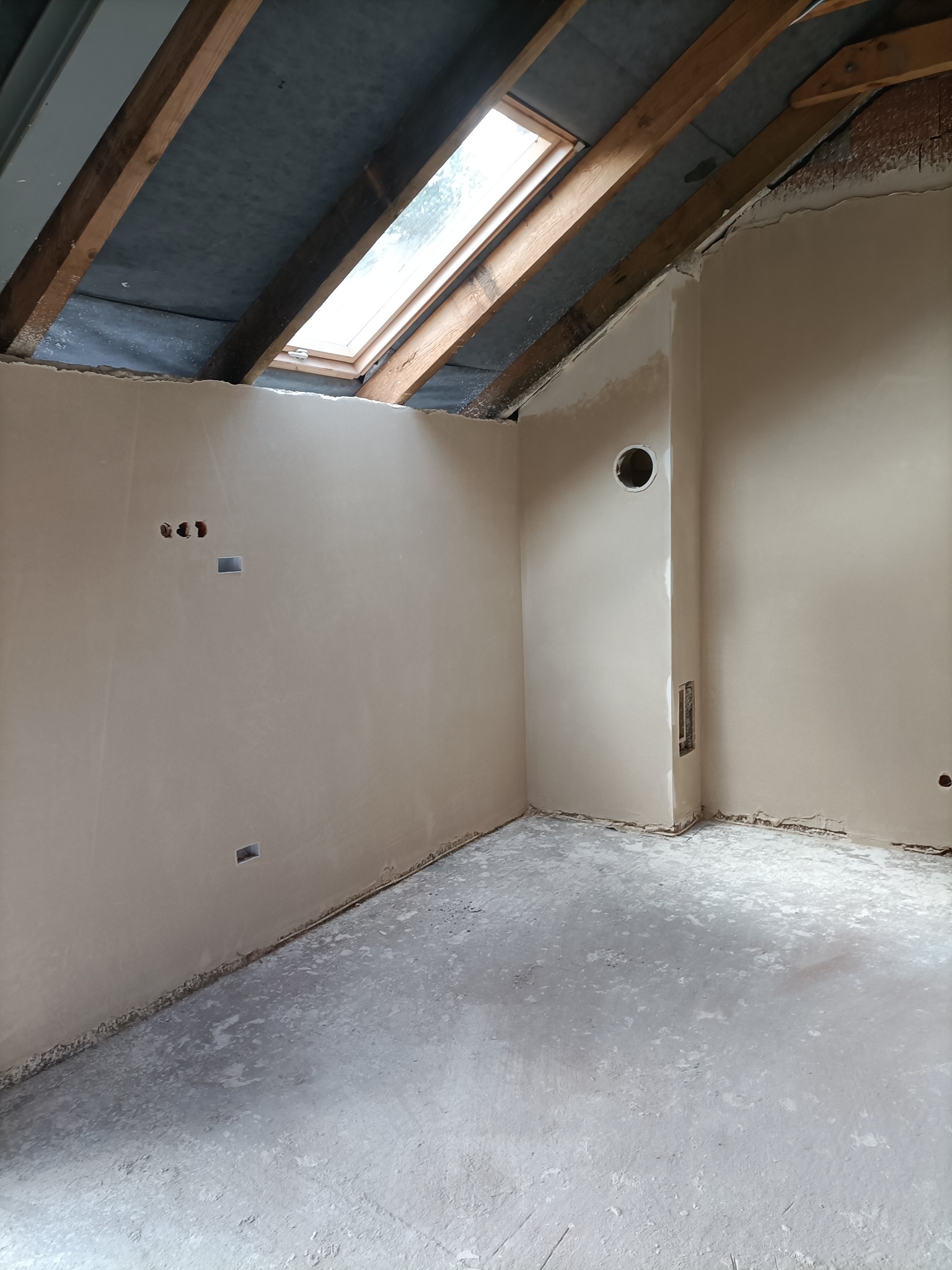 Poddasze po nałożeniu gładzi szpachlowej, widoczne otwory instalacyjne, okno dachowe i surowy strop drewniany.