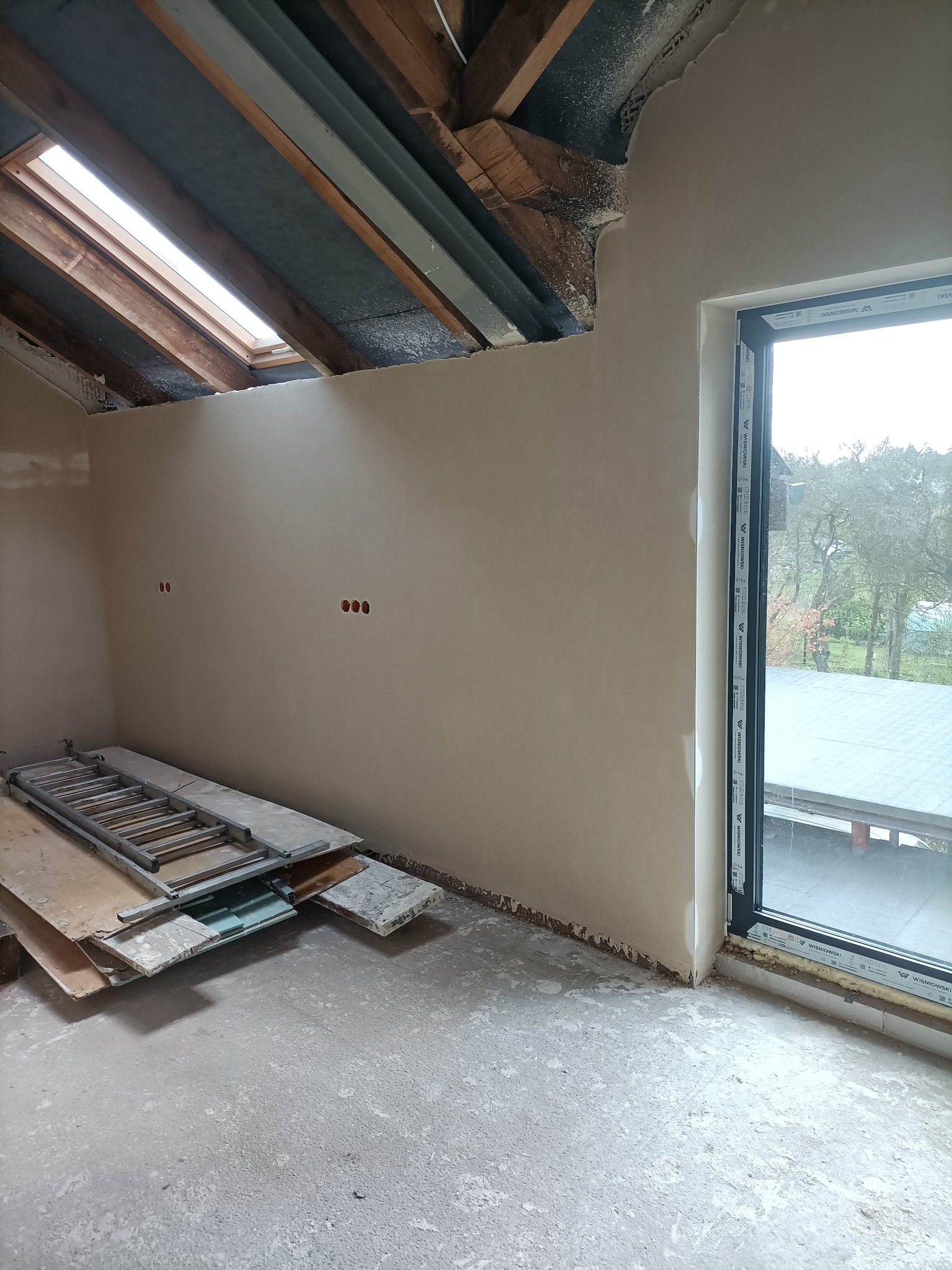 Świeżo otynkowane wnętrze poddasza z oknem dachowym, widoczne ślady prac wykończeniowych, drabina i płyty leżące na posadzce, surowe drewniane elementy konstrukcyjne dachu.